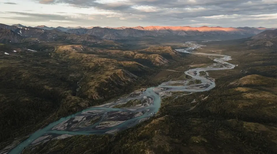 A braided turquoise river winds through dark mountain ridges under coral evening light.